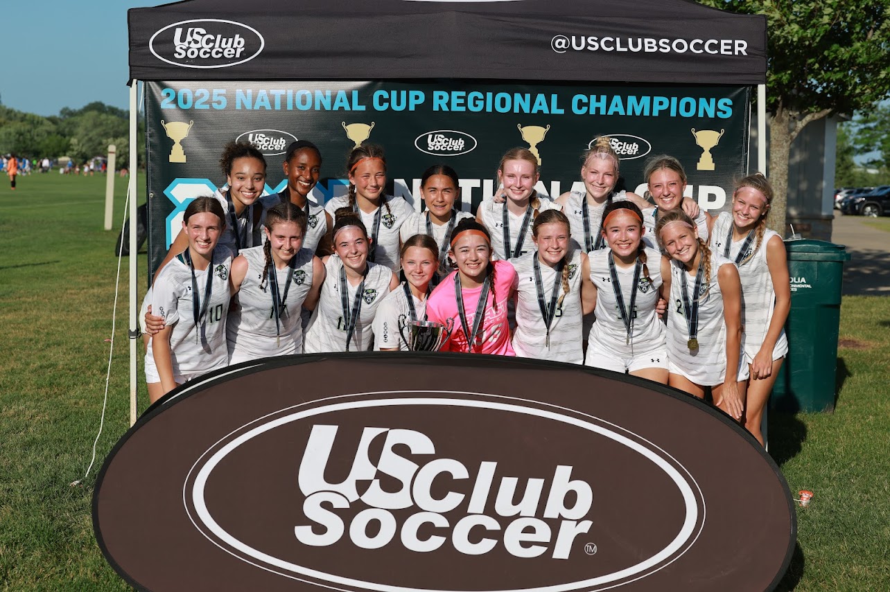 Blast Competitive team group photo, April 2026, players and coaches on turf field with trophy and soccer ball.